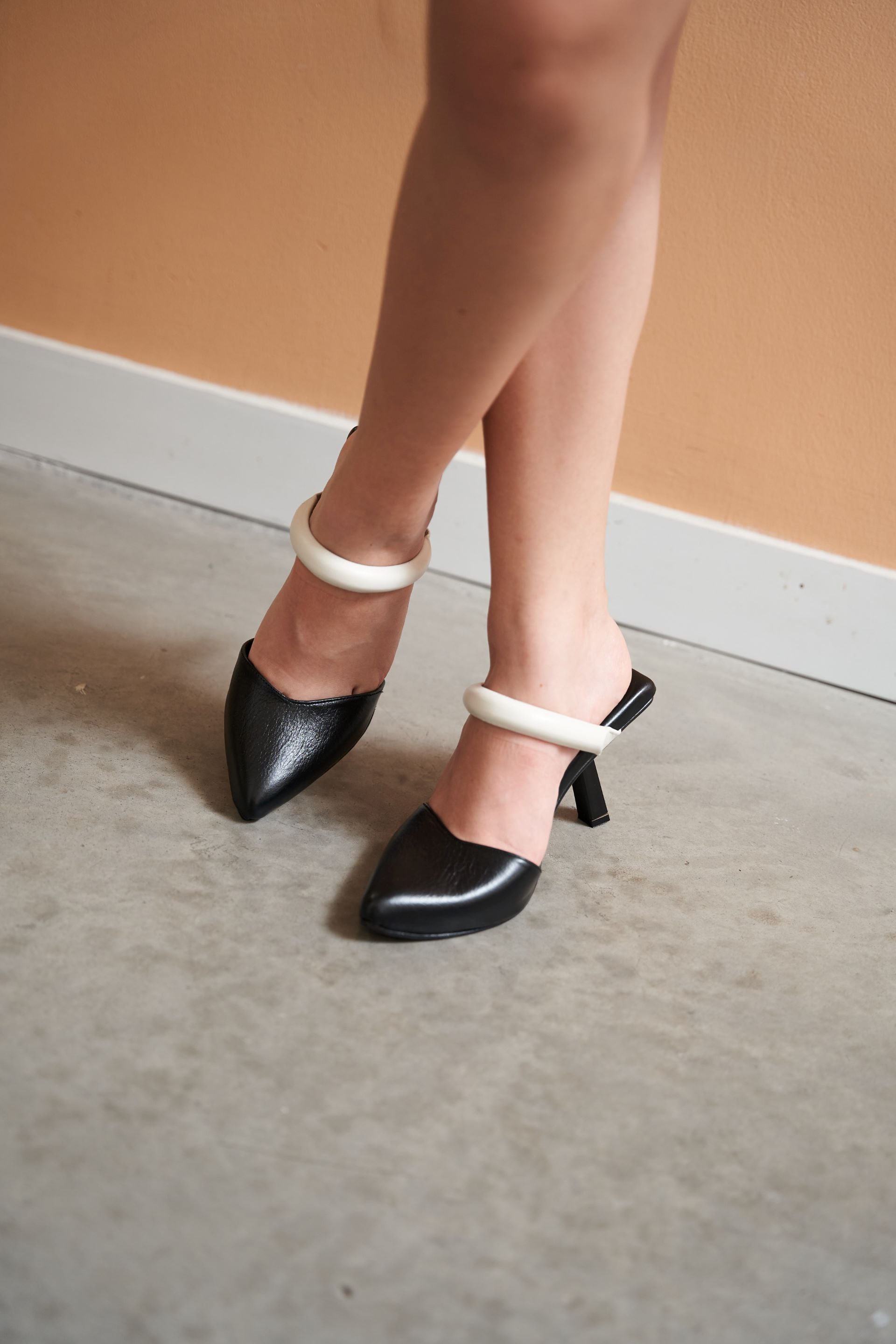 Zapatos mule de tacón 7.5CM en color negro con punta afilada y detalle de pulsera rígida en tono blanco alrededor del tobillo. Fabricados en Colombia con cuero de bovino, combinan elegancia y modernidad con un diseño único y sofisticado, ideal para realzar cualquier look.