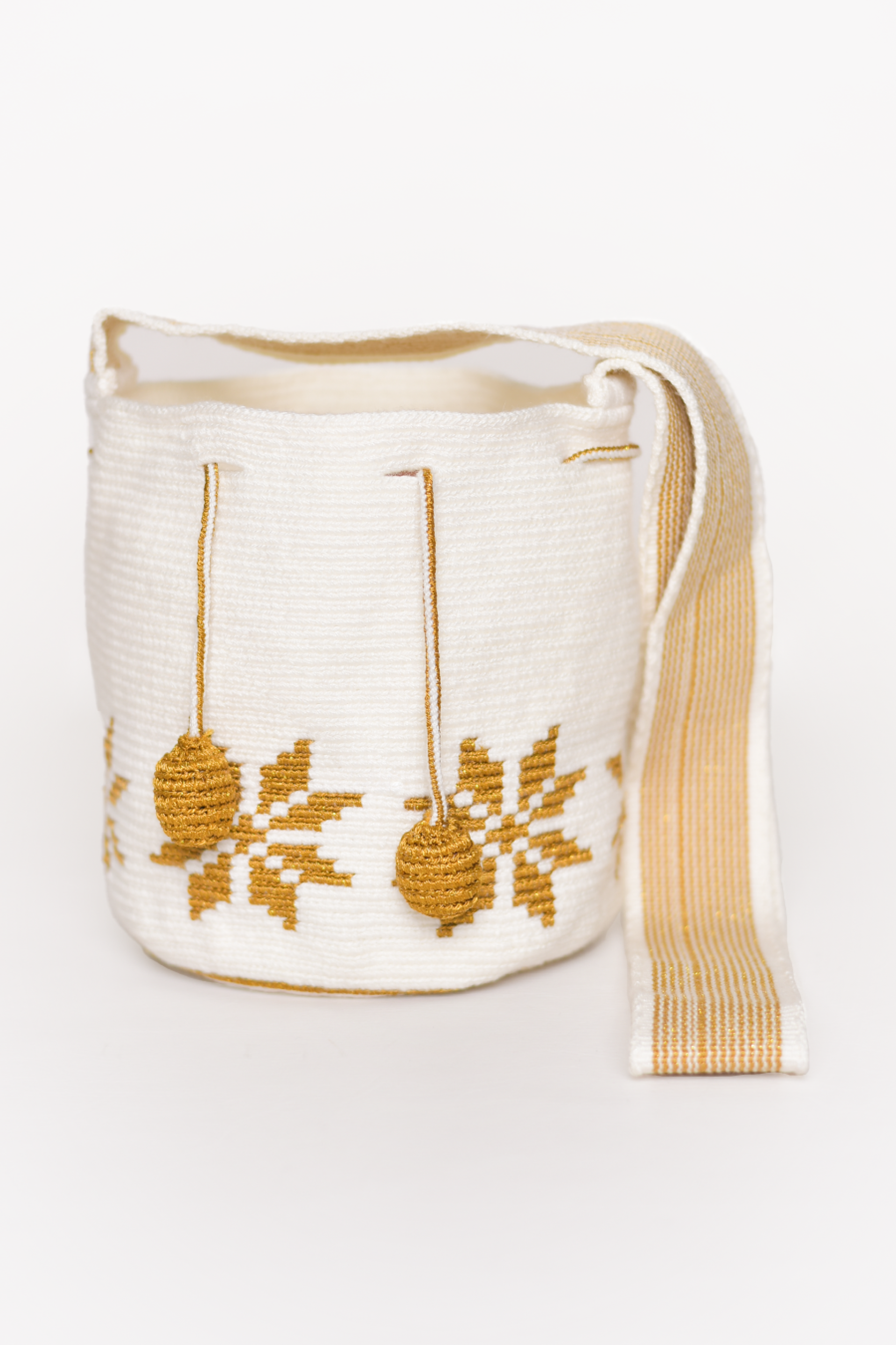 Mochila artesanal Wayuu 100% hecha a mano con hilos de algodón y seda dorada, diseñada por artesanas indígenas de la Comunidad Wayuu. Presenta un diseño de flores en tonos dorados, un asa larga, cordón ajustable y borlas decorativas, todo en color blanco. Un accesorio único que refleja la riqueza de la moda colombiana.