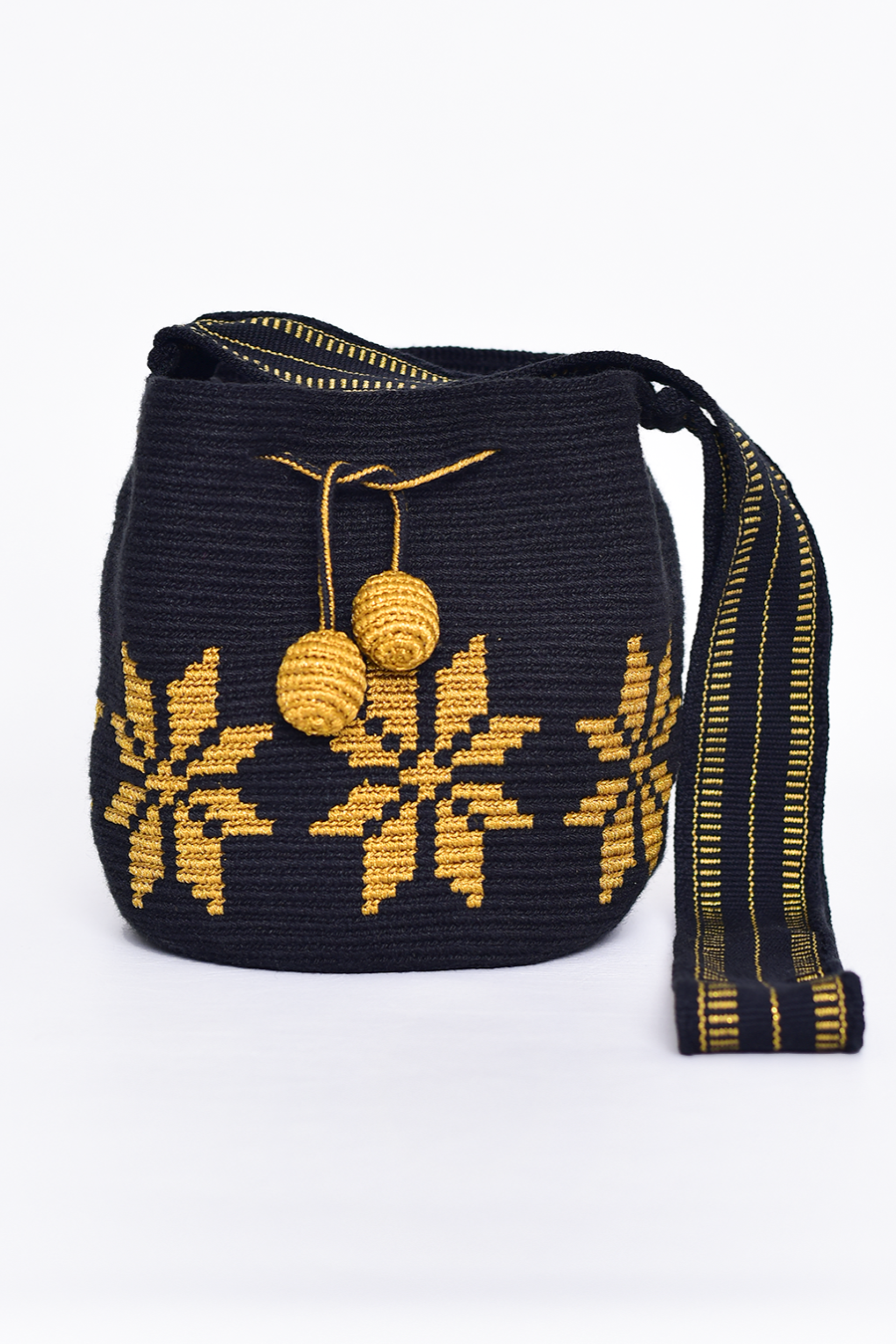 Mochila artesanal Wayuu 100% hecha a mano con hilos de algodón y seda dorada, diseñada por artesanas indígenas de la Comunidad Wayuu. Presenta un diseño de flores en tonos dorados, un asa larga, cordón ajustable y borlas decorativas, todo en color blanco. Un accesorio único que refleja la riqueza de la moda colombiana.