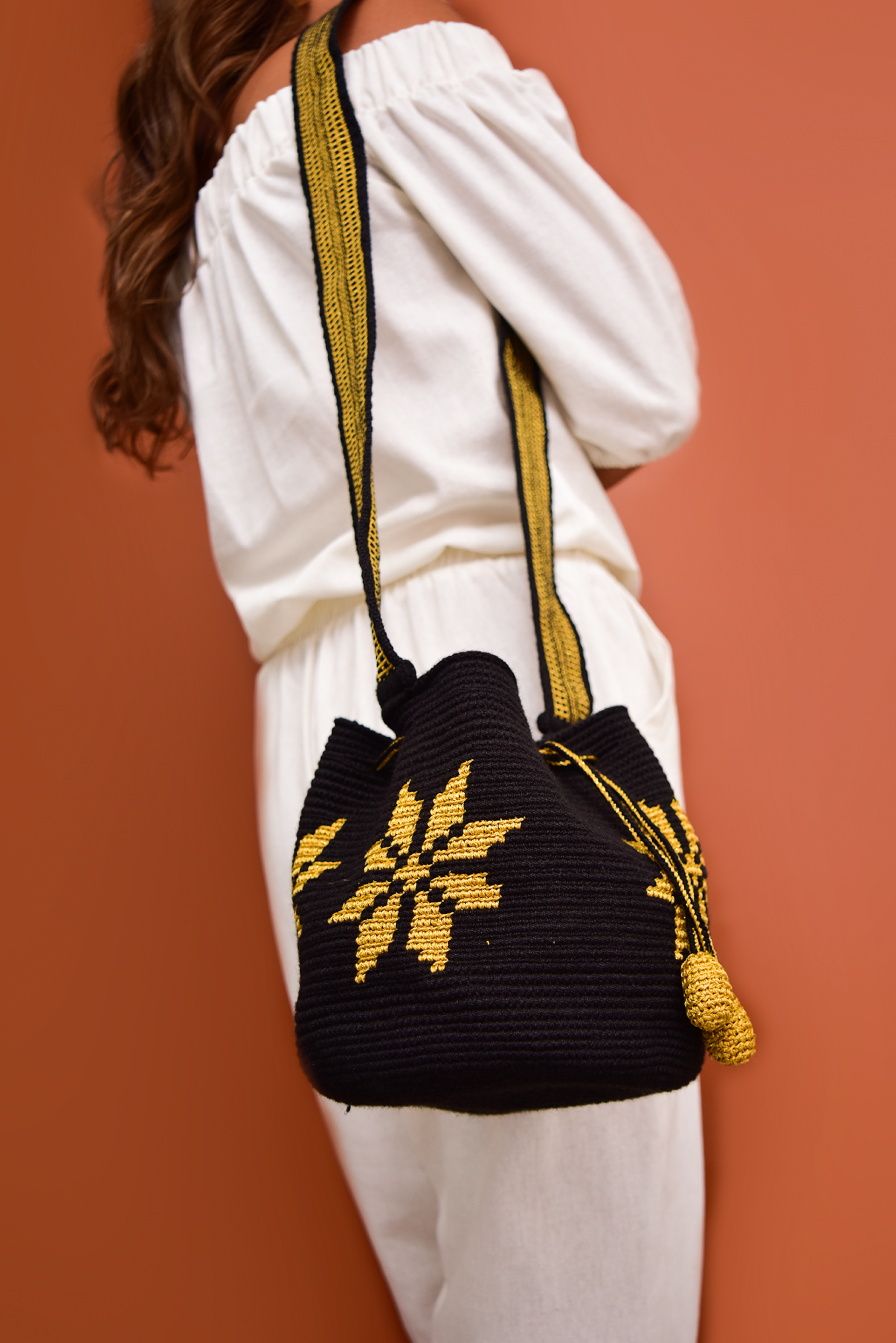 Mochila artesanal Wayuu 100% hecha a mano con hilos de algodón y seda dorada, diseñada por artesanas indígenas de la Comunidad Wayuu. Presenta un diseño de flores en tonos dorados, un asa larga, cordón ajustable y borlas decorativas, todo en color blanco. Un accesorio único que refleja la riqueza de la moda colombiana.