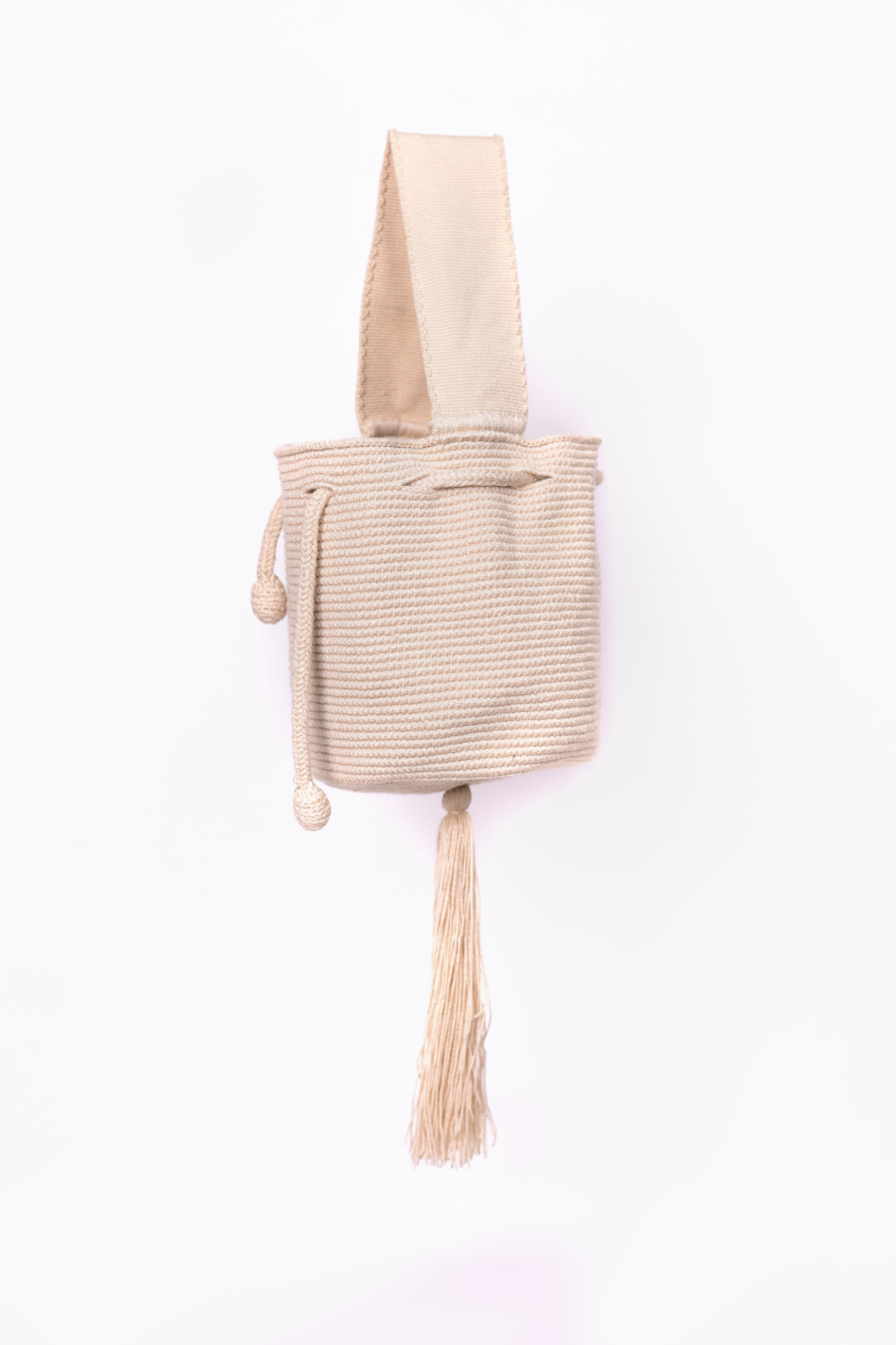 Mini mochila Wayuu artesanal, tejida a mano con hilo unicolor beige, con un colgante decorativo. Cuenta con una cargadera corta y un cordón ajustable. Hecha por artesanas de la comunidad Wayuu, reflejando la autenticidad de la moda colombiana.