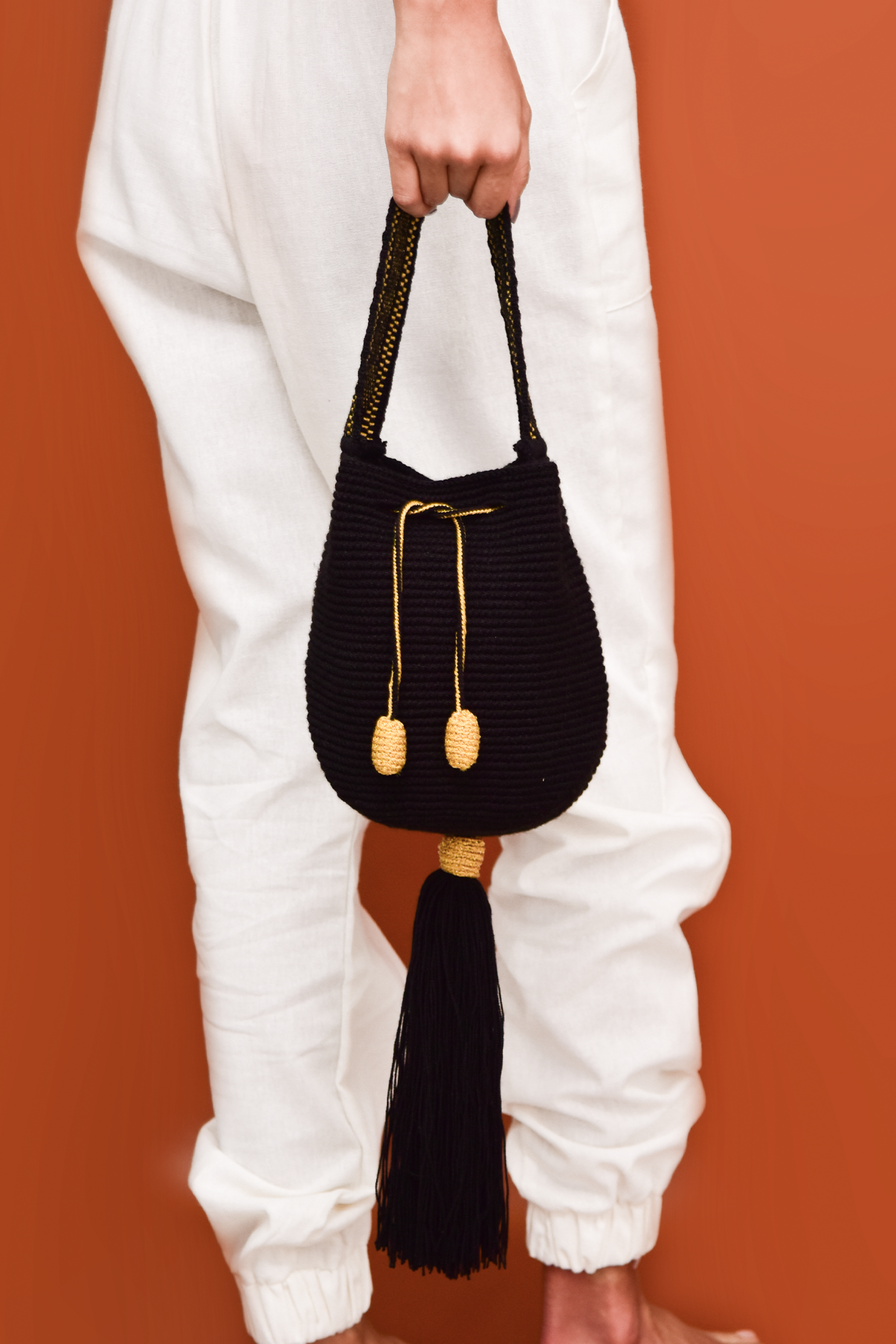 Mochila artesanal Wayuu de color negro, tejida a mano con hilos de algodón y detalles de hilos dorados. Diseño con borlas decorativas y cordón ajustable. Ideal para quienes buscan moda colombiana única y auténtica. ¡Compra online en Micerino!