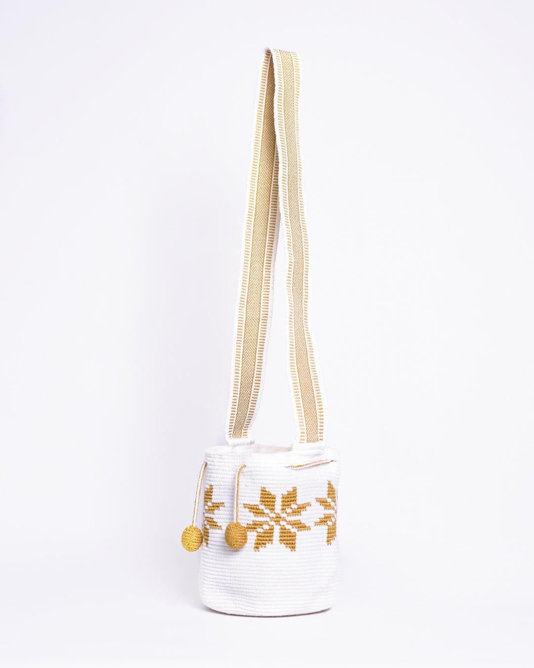 Mochila artesanal Wayuu 100% hecha a mano con hilos de algodón y seda dorada, diseñada por artesanas indígenas de la Comunidad Wayuu. Presenta un diseño de flores en tonos dorados, un asa larga, cordón ajustable y borlas decorativas, todo en color blanco. Un accesorio único que refleja la riqueza de la moda colombiana.