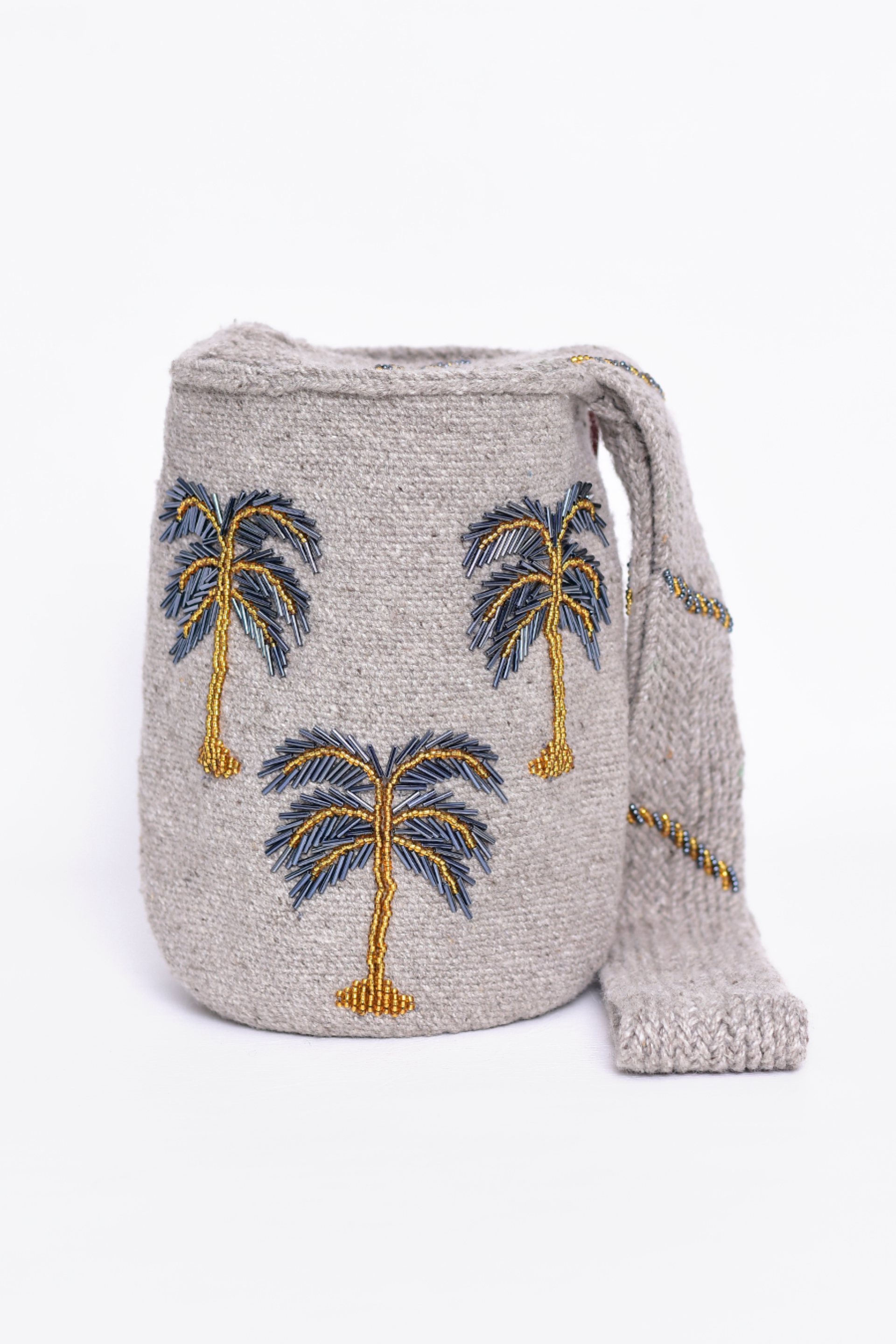 Mochila Kankuama hecha a mano, con bordado detallado de palmeras en colores grises que evocan la esencia tropical. La mochila, confeccionada en material de alta calidad, presenta una correa tejida que resalta el trabajo artesanal de la comunidad Kankuama en Colombia. Un accesorio único y auténtico de moda colombiana